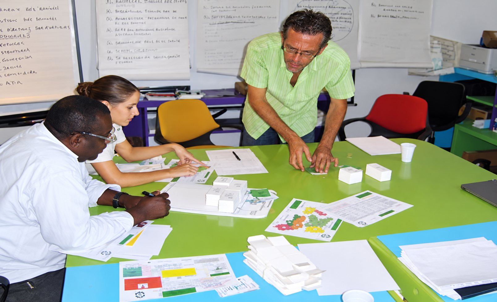 Trois personnes travaillent autour d'une grande table verte couverte de plans, de documents et de petites maquettes blanches dans une salle de réunion.