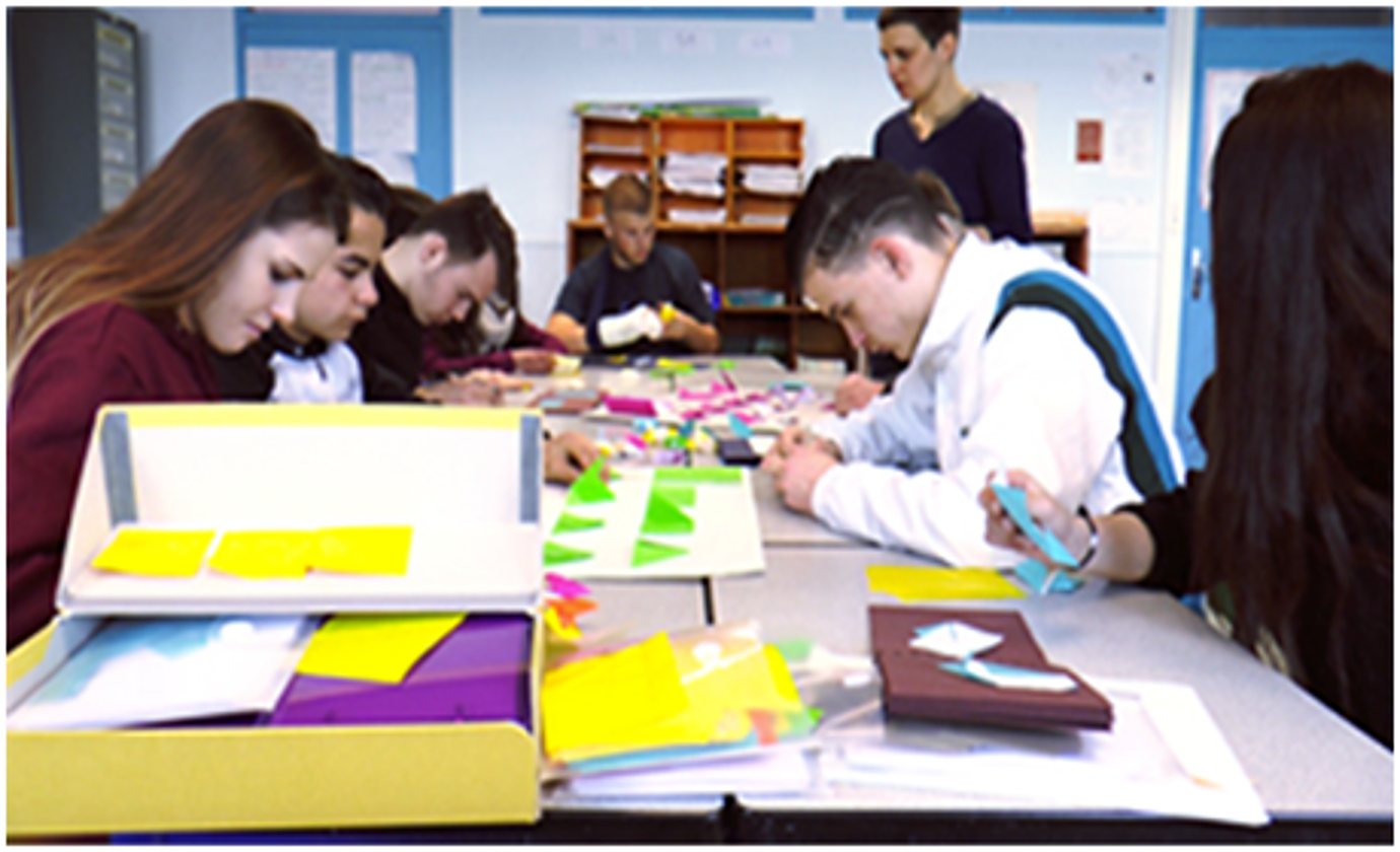 Des adolescents assis autour de grandes tables manipulent des pièces de papier et de carton colorées dans une salle de classe, avec une enseignante debout au fond.