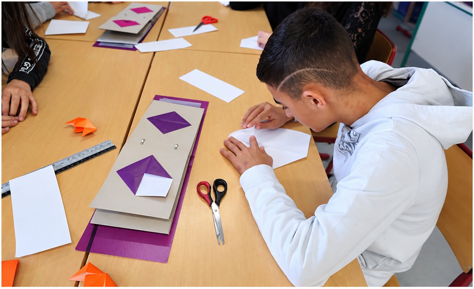Un jeune garçon en sweat blanc plie une feuille de papier à une table en bois où sont posés des ciseaux, une règle et des modèles en carton violet.