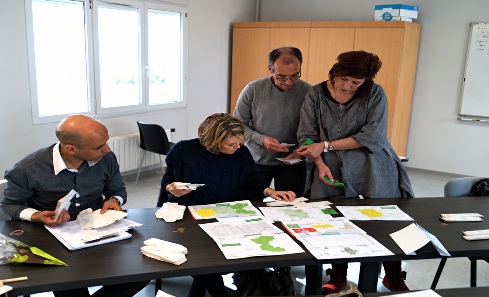 Quatre personnes, deux assises et deux debout, examinent des feuilles imprimées colorées posées sur de grandes tables dans une salle lumineuse.