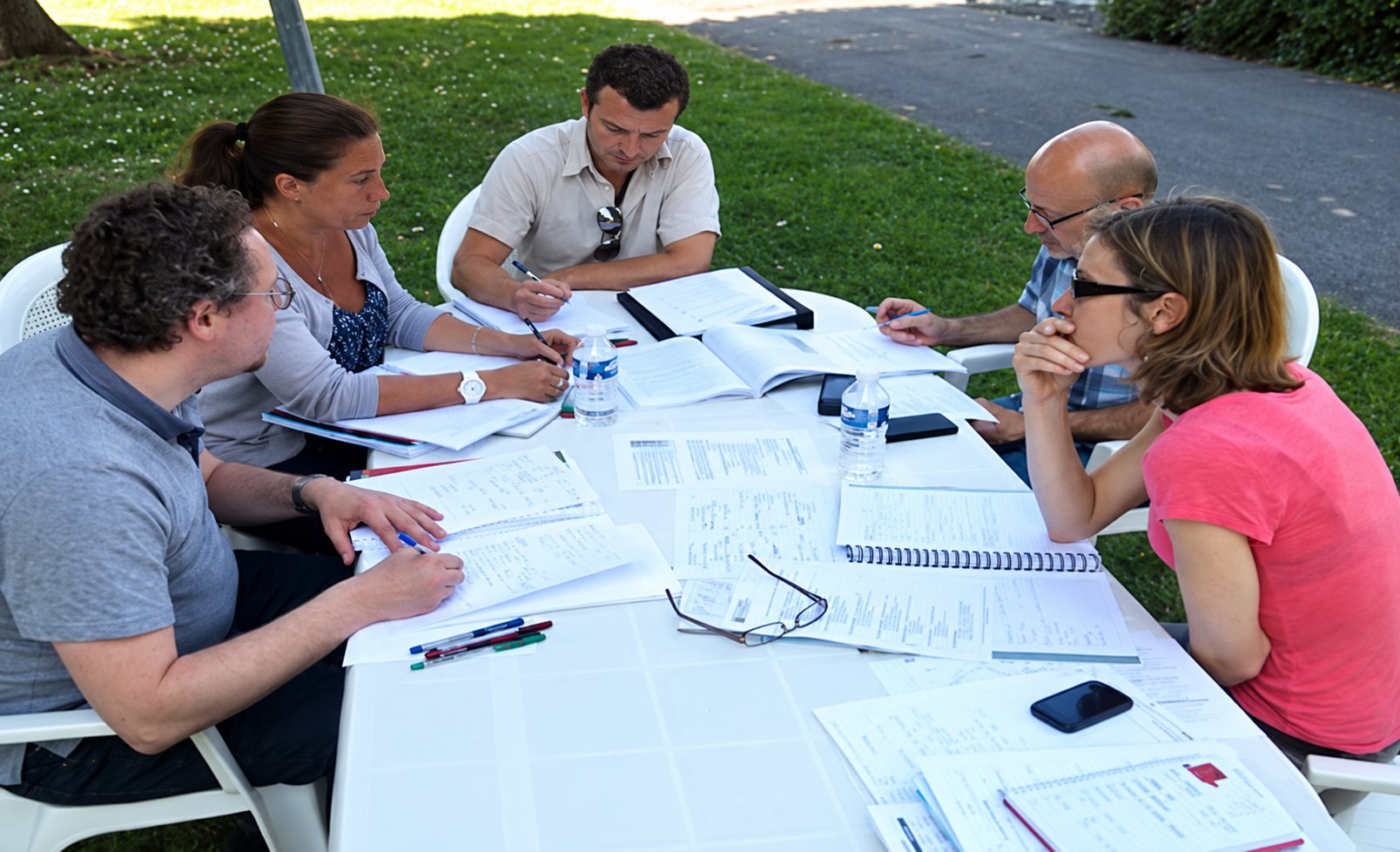 Six personnes assises autour d'une table blanche en plein air travaillent sur des documents et des cahiers posés devant elles.