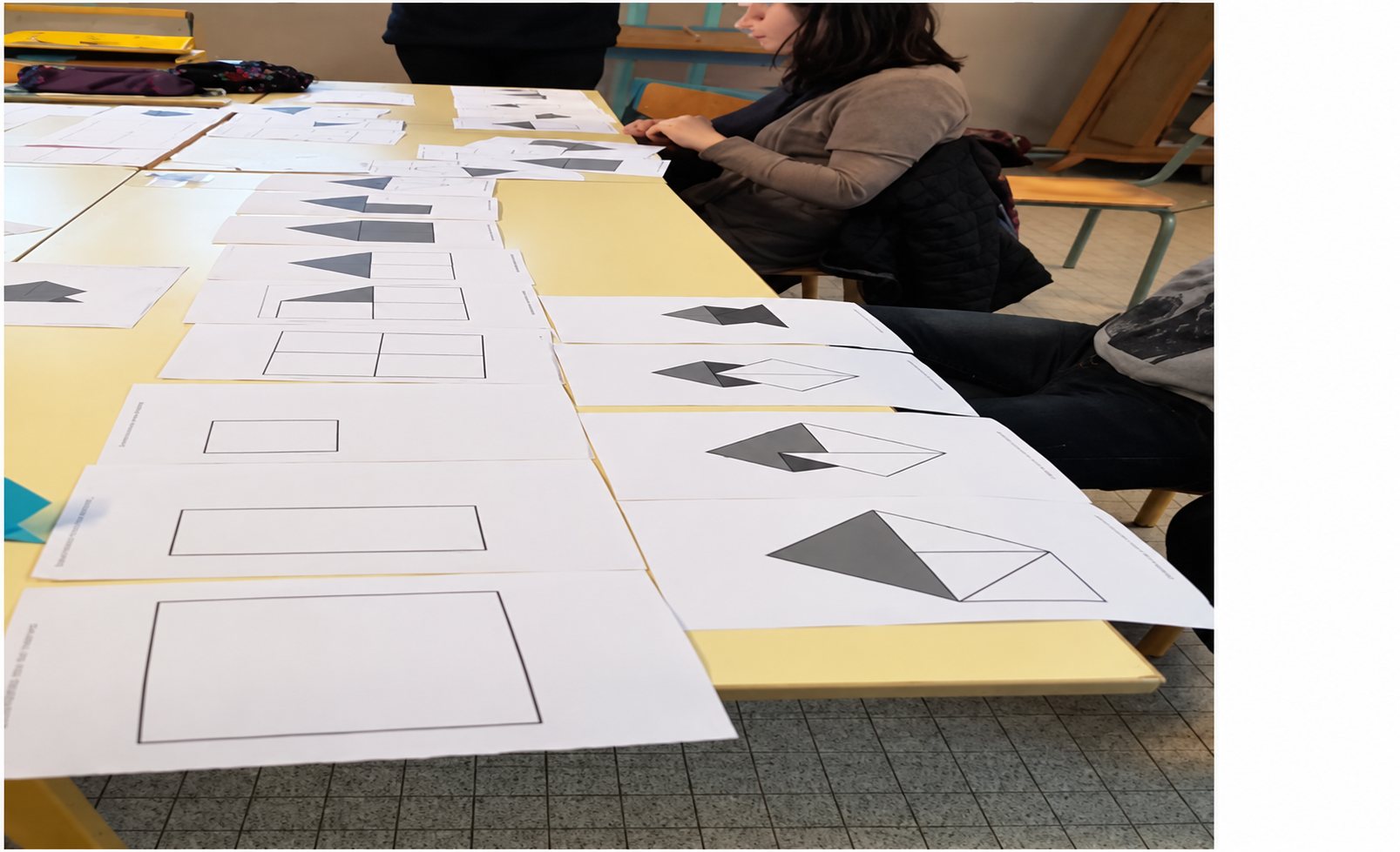 Des feuilles blanches imprimées de rectangles et de triangles gris sont alignées sur des tables jaunes, avec des personnes assises à l'arrière-plan.