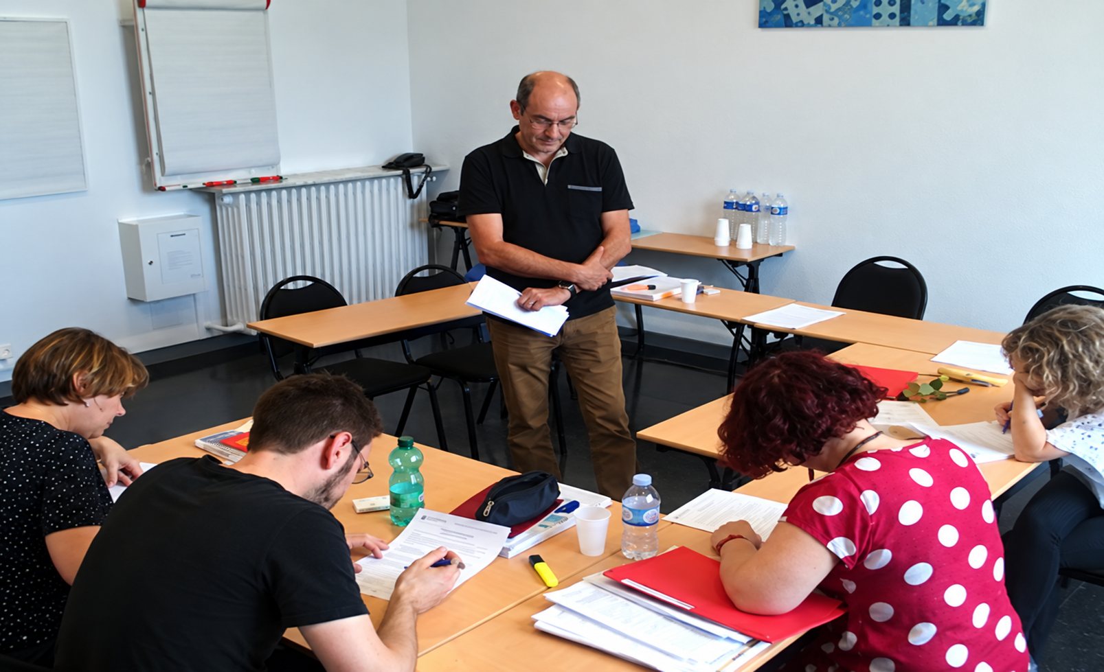 Un formateur tenant des feuilles se tient debout dans une salle de classe devant plusieurs stagiaires assis à des tables qui prennent des notes.