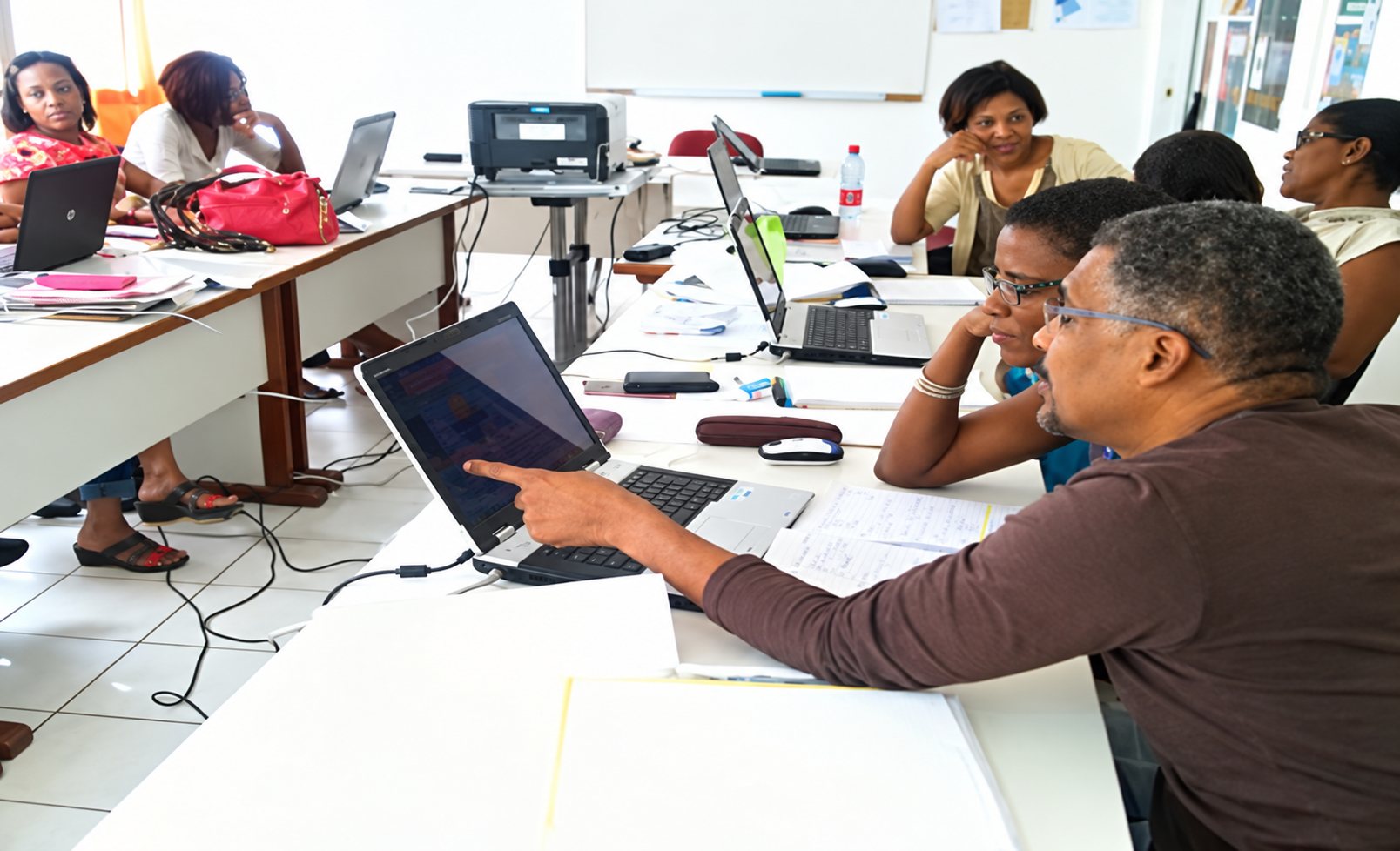 Un formateur montre l'écran d'un ordinateur portable à un participant dans une salle de formation où d'autres personnes travaillent aux tables.
