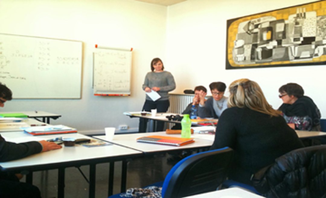 Dans une salle de classe lumineuse, une formatrice se tient debout devant des adultes assis à des tables, avec des tableaux blancs couverts de notes derrière elle.