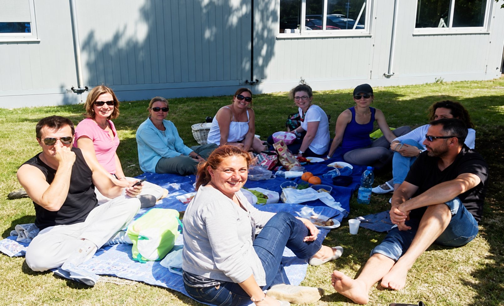 Un groupe de huit personnes est assis sur une couverture dans l'herbe et partage un pique-nique devant un bâtiment gris par une journée ensoleillée.