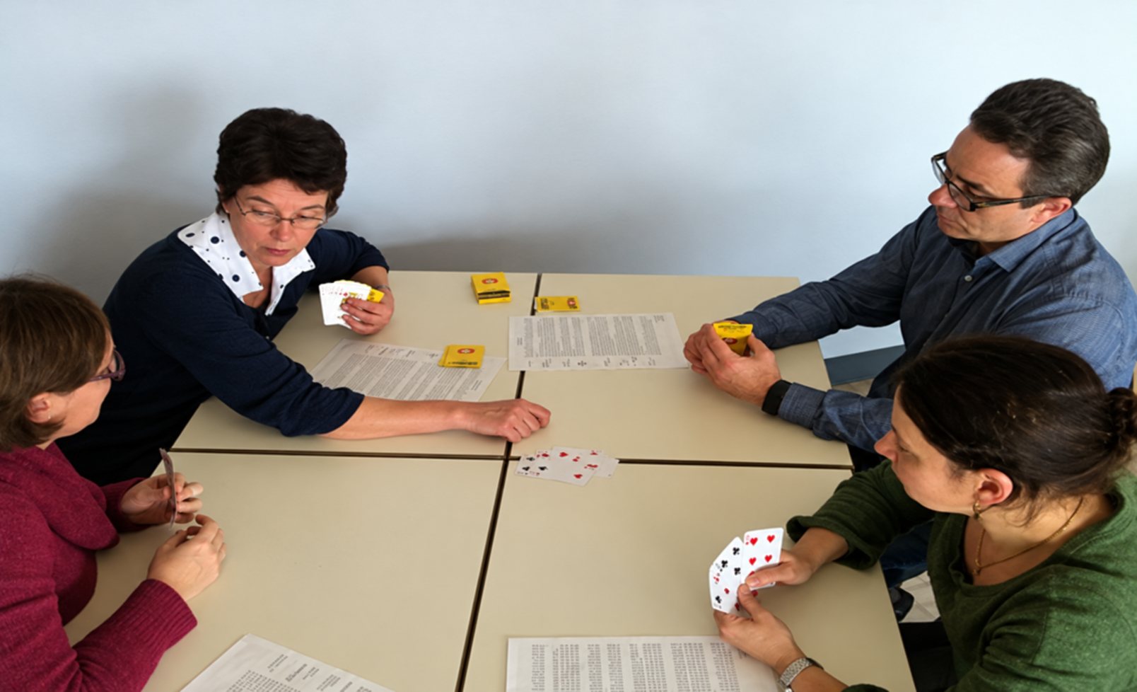 Quatre personnes sont assises autour d'une table et jouent aux cartes, avec des feuilles de papier et de petits objets jaunes posés devant elles.