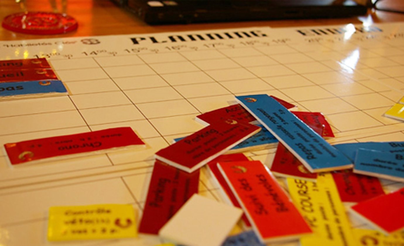 Un grand planning blanc quadrillé sur une table, recouvert d'étiquettes rouges, bleues et jaunes portant des inscriptions manuscrites.