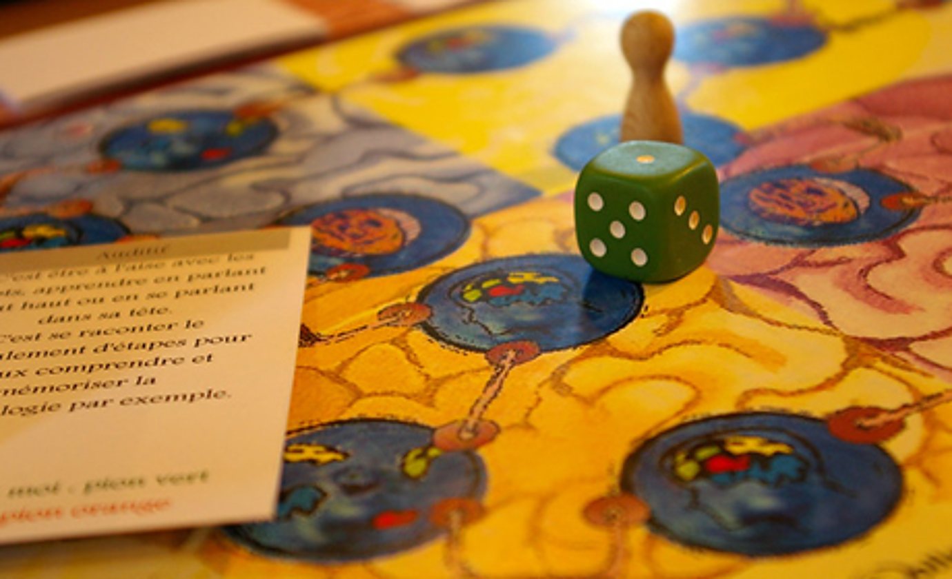 Gros plan sur un plateau de jeu illustré de cerveaux colorés, avec un dé vert, un pion en bois et une carte de règles au premier plan.