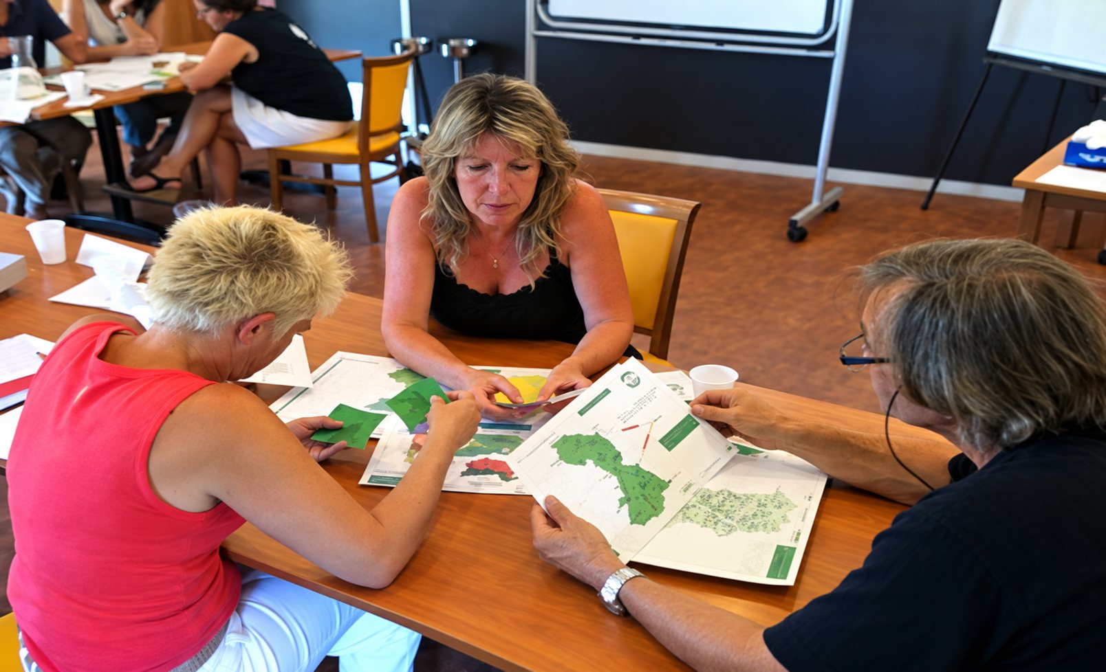 Trois personnes sont assises autour d'une table en bois et examinent ensemble des cartes et des documents imprimés dans une salle de réunion.