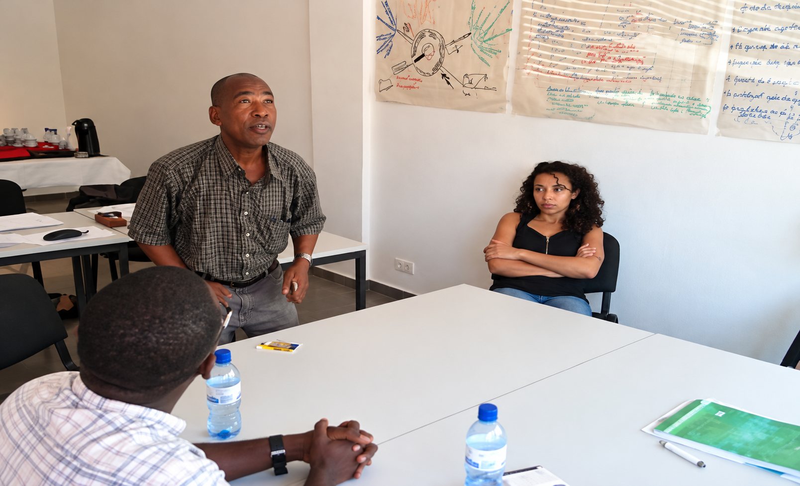 Dans une salle de réunion, un homme debout prend la parole devant une femme assise, des bouteilles d'eau posées sur les tables et des affiches au mur.
