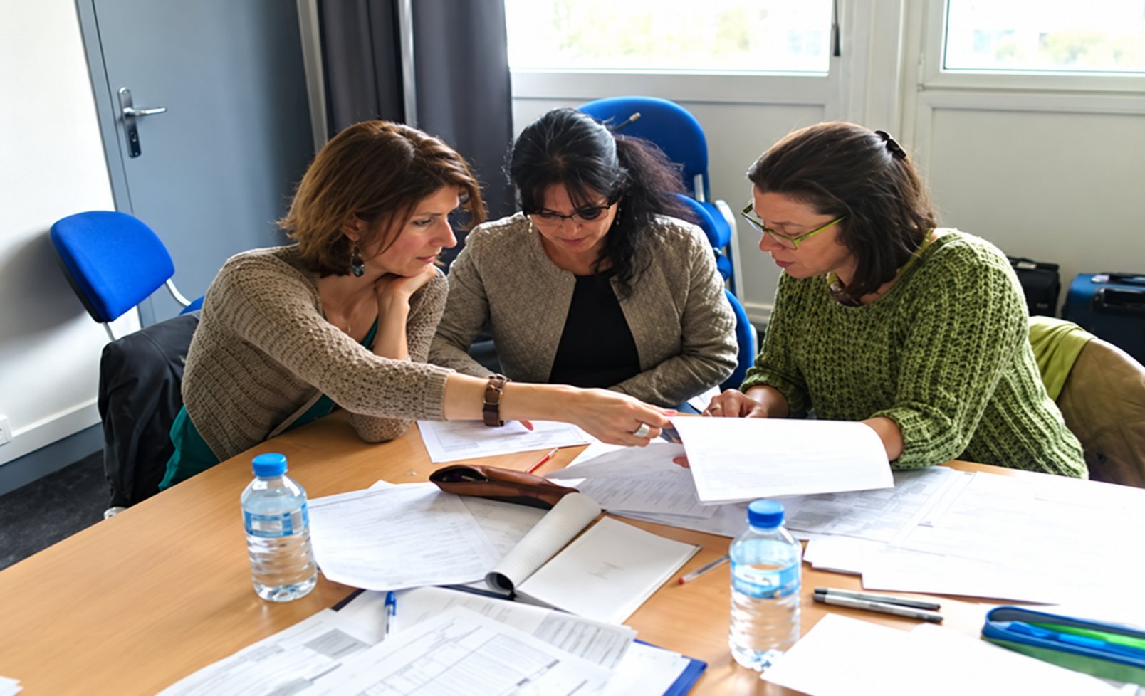 Trois femmes sont assises autour d'une table en bois et examinent ensemble des documents, avec des bouteilles d'eau posées devant elles.